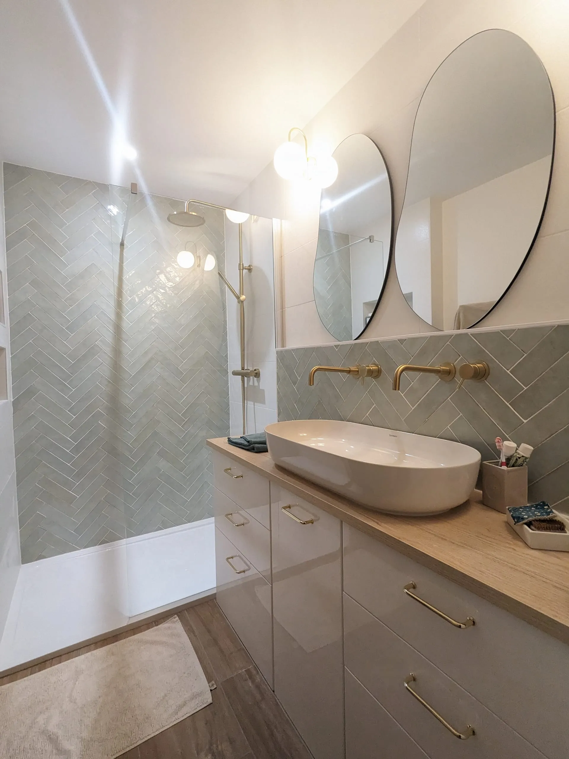Salle de bain terminée avec douche ouverte, carrelage gris en chevrons, meuble en bois et robinetterie en laiton